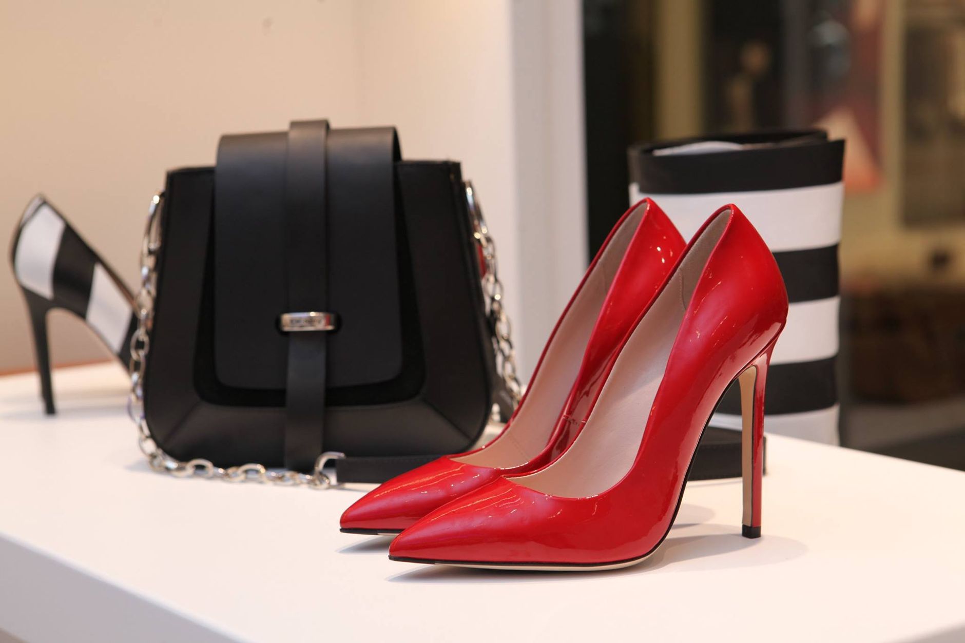 Display of red patent leather heels, black shoulder bag with chain strap, and striped cylindrical container on white surface