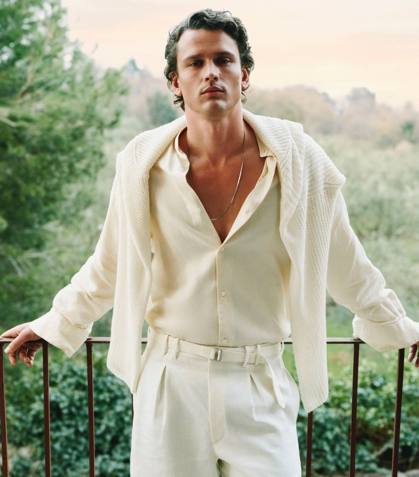 Man in cream linen suit and cardigan leaning on railing overlooking green landscape