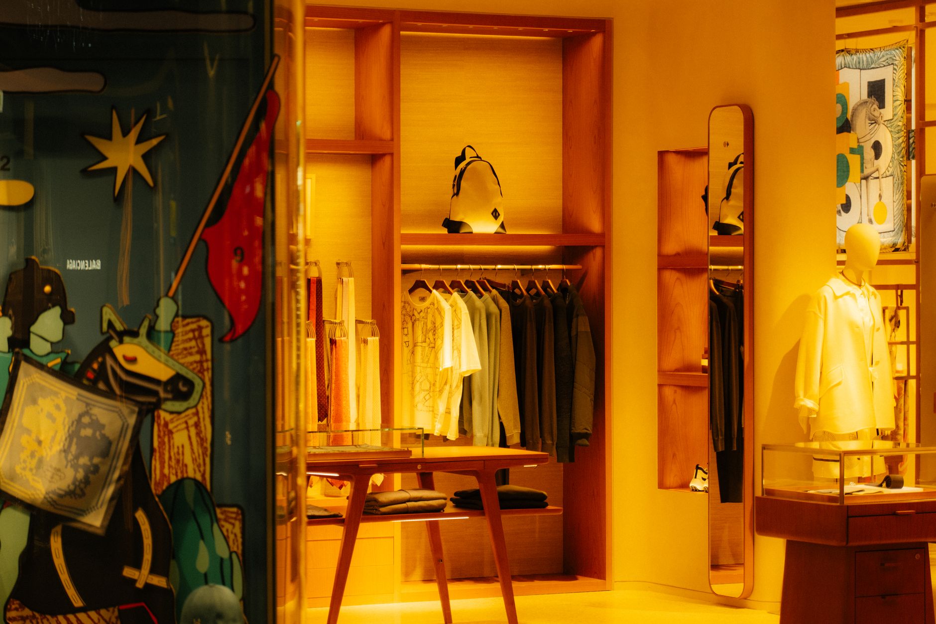Warm-lit boutique interior with orange walls, clothing racks displaying apparel, mannequin in yellow shirt, and decorative artwork